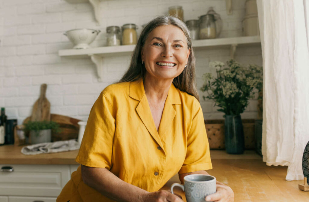 A senior woman with long gray hair looking directly smiling and holding a cup of tea.