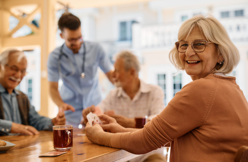 A senior woman sits at a table, holding cards next to a cup of tea and smiling. In the background, fellow seniors engage in conversation while a nurse helps out.
