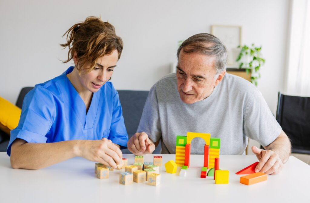 A caregiver in a blue uniform assists an older adult in building colorful block structures at a table to help stimulate cognitive abilities for dementia care.