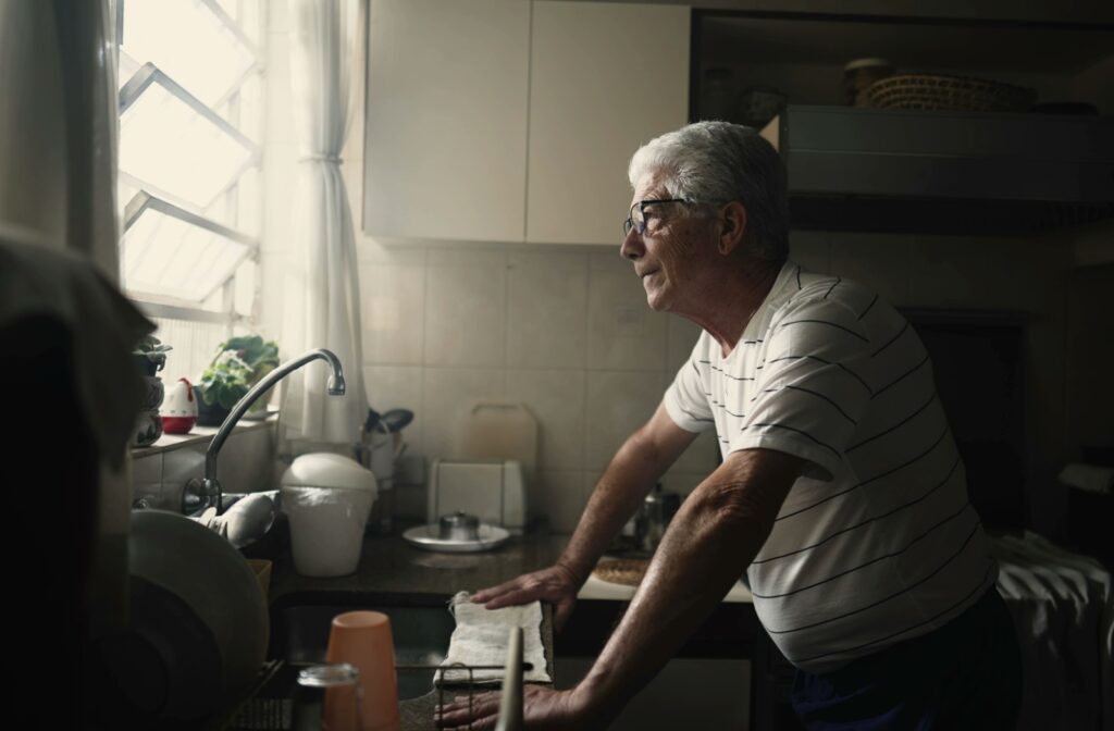 A senior rests against the kitchen counter, looking out the window, while struggling with the task of keeping the house clean